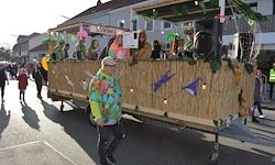 Ein bunter Wagen mit Leuten darauf bewegt sich die Straße hinunter. Ein Mann in einem bunten Hemd geht neben ihm her.