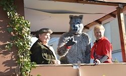 Zwei Menschen stehen auf einem Balkon, einer ist als Wolf verkleidet, während ein Mann in einem Hut in der Nähe steht.