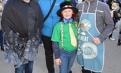 Drei Personen in verschiedenen Kostümen stehen dicht beieinander, eine hält ein Schild mit den Worten 'Fresh Meat Burgers.' Sie scheinen sich bei einem Umzug zu befinden.