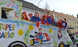 Ein dekorierter Aufzug mit Menschen im Inneren, der ein Super-Mario-Thema zeigt, bewegt sich in einer Parade. Der Aufzug hat farbenfrohe Zeichnungen, und die Menschen tragen passende Kostüme.