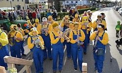 Eine als Minions verkleidete Marschkapelle geht die Straße entlang, während eine Menschenmenge zuschaut. Einige tragen Musikinstrumente, und einer trägt eine Handtasche.