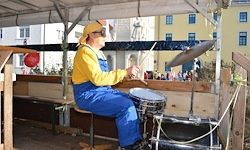 Ein Mann in gelber und blauer Arbeitskleidung spielt Trommeln und Becken im Freien. Er sitzt auf einem Hocker, während eine Menge Menschen im Hintergrund zuschaut.