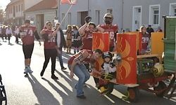 Eine Gruppe von Menschen in roten Trikots mit Nummern zieht auf einer Straße. Ein Mann schiebt einen Wagen mit einem Kind und einem Baby. Gebäude säumen die Straße, Schatten fallen auf den Boden.