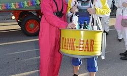 Eine Person in einem rosa Overall hält einen gelben Eimer mit der Aufschrift Tank-Bar, während eine andere Person in einer gelben Jacke und blauer Hose neben ihr steht. Im Hintergrund ein geschmücktes Fahrzeug mit einem roten Rad und mehreren Personen in Kostümen.