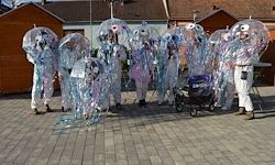 Eine Gruppe von Menschen in Quallenkostümen und weißen Anzügen steht in einer Reihe, mit einem Kinderwagen und Häusern im Hintergrund.