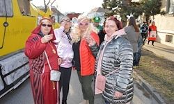 Vier Frauen stehen gemeinsam auf einer Straße, gekleidet in festliche Kostüme. Hinter ihnen ist ein großes gelbes Fahrzeug geparkt. Die Frauen tragen Sonnenbrillen und halten verschiedene Gegenstände. Im Hintergrund sind Menschen und Gebäude zu sehen.