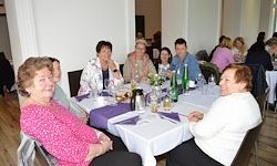 Eine Gruppe von Menschen sitzt in einem Restaurant um einen Tisch. Der Tisch hat Blumen, Flaschen und Gläser. Der Raum hat Holzböden und weiße Wände.
