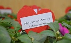 Ein rotes herzförmiges Kärtchen mit der Botschaft 'Alles Liebe zum Muttertag vom Bürgermeister' steht in einem Garten mit grünen Blättern und einer rosa Blume.