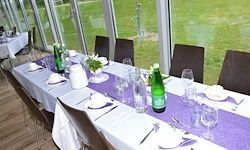 Ein langer Tisch ist für eine formelle Veranstaltung gedeckt mit weißer Tischdecke, lila Tischläufern, Gläsern, Tellern, Tassen und zwei grünen Flaschen. Der Raum hat große Fenster mit Blick auf ein saftiges Feld.