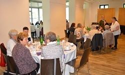 Eine Gruppe älterer Frauen sitzt um einen Tisch in einem Speisesaal. Sie essen. Es gibt Flaschen und Teller auf dem Tisch. Eine Person steht hinter ihnen.