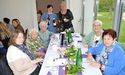 Mehrere Frauen sitzen an einem langen Tisch mit Flaschen und Blumen, wahrscheinlich bei einer sozialen Veranstaltung oder Zusammenkunft. Sie lächeln und scheinen sich zu amüsieren.