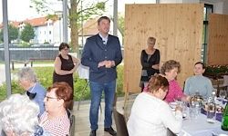 Ein Mann hält eine Rede bei einer Versammlung. Er steht vor einer Gruppe von Menschen, die an einem Tisch sitzen. Einige tragen Brillen und einige halten Weinflaschen. Dahinter ist eine Holzwand zu sehen.