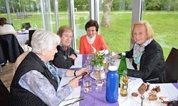 Vier ältere Frauen sitzen in einem Restaurant an einem Tisch mit Blick auf ein Grasfeld. Auf dem Tisch stehen Teller, Gläser, eine Blumenvase und Flaschen.