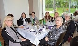 Eine Gruppe von Frauen mit Brillen und Schals sitzt um einen Tisch mit Flaschen und Gläsern. Sie genießen wahrscheinlich gemeinsam eine Mahlzeit im Esszimmer.
