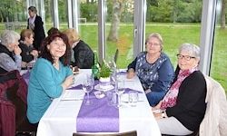 Eine Gruppe von Frauen sitzt an einem Tisch im Inneren eines Restaurants, mit Blick auf eine Glaswand mit einer Wiese und Bäumen.