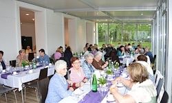 Eine große Gruppe von Menschen sitzt in einem Restaurant an Tischen, mit Gläsern, Flaschen und Pflanzen auf den Tischen.
