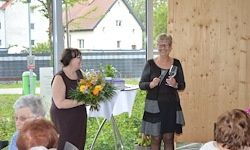 Zwei Frauen stehen in einem Raum, eine hält Blumen und die andere eine Trophäe. Ein Publikum sitzt hinter ihnen. Ein Tisch mit einer weißen Tischdecke steht vor ihnen. Eine Holzwand und ein Haus sind im Hintergrund.