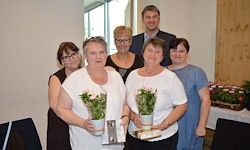 Eine Gruppe von Frauen mit Blumen und Auszeichnungen steht zusammen und lächelt für ein Foto. Ein Tisch mit Blumen ist im Hintergrund.