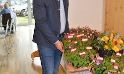 Ein Mann steht vor einem Tisch mit Blumenarrangements, mit Menschen, die am Tisch im Hintergrund sitzen.