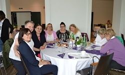 Eine Gruppe von Menschen sitzt an einem runden Tisch mit einer weißen Tischdecke, Tellern und Flaschen. Sie scheinen eine Mahlzeit einzunehmen. Einige tragen Brillen. Im Hintergrund befindet sich eine Tafel, und eine Person steht in der Nähe der Wand.