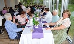 Mehrere ältere Frauen sitzen an einem langen Tisch mit weißen Tischdecken und lila Tischläufern, möglicherweise in einem Restaurant. Auf dem Tisch stehen Topfpflanzen und Weinflaschen. Der Raum hat große Glasfenster.