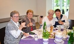 Vier ältere Frauen sitzen um einen Tisch herum, halten Weingläser in den Händen und scheinen einen besonderen Anlass zu feiern. Der Tisch ist mit Weinflaschen, einer Topfpflanze und einem Kuchen gedeckt.