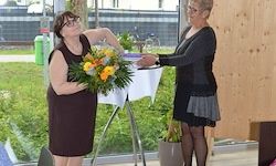 Zwei Frauen stehen bei einem Tisch mit einem Blumenstrauß. Die Frau links trägt ein braunes Kleid und eine Brille, die Frau rechts trägt ein schwarzes Kleid. Ein Topf mit einer Pflanze steht rechts.