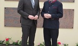 Zwei Männer in Anzügen stehen nebeneinander vor einer Wand mit Paneelen und Blumen.