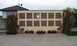 Eine große, beige Wand mit mehreren rechteckigen Platten, möglicherweise Gedenktafeln, steht in einem Garten mit mehreren Pflanzen und Blumen davor.