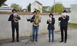 Vier Musiker spielen im Freien Blechblasinstrumente und stehen in einer Reihe vor einer Wand. Sie tragen alle Hüte und schwarze Schuhe.