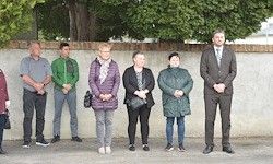 Eine Gruppe von sechs Personen steht vor einer weißen Wand, wahrscheinlich an einer Zeremonie teilnehmend. Sie tragen alle formelle Kleidung.