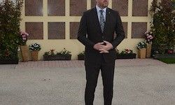 Ein Mann in einem Anzug steht mit verschränkten Armen vor einer Gedenkwand, umgeben von Blumen und Pflanzen.
