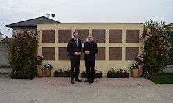 Zwei Männer stehen vor einer gelben Wand mit Gedenktafeln, umgeben von Pflanzen und Blumen. Im Hintergrund befindet sich ein Haus mit Schornstein und bewölktem Himmel.