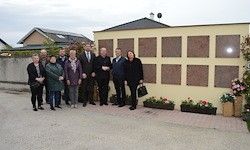Eine Gruppe von Menschen steht vor einer Gedenkwand mit Blumen, schick gekleidet, an einem bewölkten Tag.