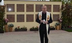 Ein Mann steht vor einer Wand mit mehreren Gedenktafeln und hält ein Stück Papier in der Hand. Der Bereich ist mit verschiedenen Topfpflanzen und Blumen geschmückt.
