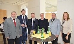 Eine Gruppe von formell gekleideten Personen steht um einen Tisch mit Getränken in Gläsern und Flaschen. Sie scheinen sich bei einer formellen Veranstaltung zu befinden.