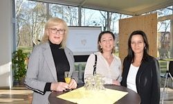 Drei Frauen stehen neben einem Tisch mit Gläsern und einer gelben Tischdecke vor einer Glaswand mit Blick auf Bäume und eine Projektionsleinwand.