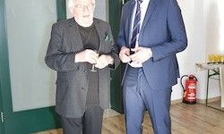 Zwei Männer stehen in einem Raum; einer hält ein Glas, der andere ein kleines Objekt. Ein Feuerlöscher und ein Tisch mit Getränken sind im Hintergrund.