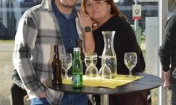 Zwei Personen, ein Mann und eine Frau, stehen hinter einem kleinen Tisch mit Flaschen, Gläsern und einer Karaffe. Sie lächeln beide und scheinen für ein Foto zu posieren.
