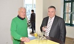 Zwei Männer stehen an einem Tisch in einem Raum mit Gläsern und Weingläsern. Ein Mann hält ein Glas und der andere trägt eine Krawatte. Ein Mann im Anzug steht hinter ihnen.