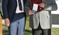 Zwei Männer stehen nebeneinander, einer hält eine Medaille und der andere trägt eine Krawatte.