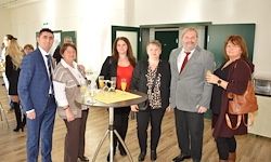 Eine Gruppe von Menschen in formeller Kleidung steht um einen Tisch mit Getränken. Dahinter befinden sich Türen und eine weiße Wand. Eine Person hält ein gelbes Papier.