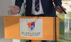 Eine Person spricht an einem Rednerpult mit dem Schild 'Schwadorf' vor einer Glaswand mit sichtbarem grünem Rasen.
