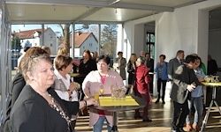 Eine Gruppe von Menschen trifft sich drinnen, möglicherweise bei einer gesellschaftlichen Veranstaltung. Einige stehen, andere sitzen. Eine Frau mit Brille hält ein Glas, und ein Tisch vor ihr hat ein gelbes Tablett.