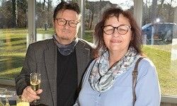 Ein Mann und eine Frau stehen nah beieinander, lächelnd, ein Glas Champagner haltend. Sie sind in einem Raum mit großen Fenstern, die auf eine Rasenfläche und ein Auto blicken.