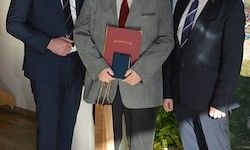 Drei Männer in Anzügen stehen zusammen, einer hält ein rotes Buch und ein blaues Objekt in den Händen.