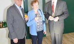 Drei Menschen in formeller Kleidung stehen mit Champagner-Gläsern in der Hand in einem Raum mit Holzboden und grünen Türen.