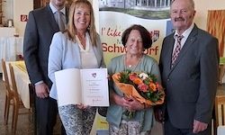 Drei Menschen stehen eng beieinander, zwei Frauen und ein Mann, alle formell gekleidet. Eine Frau in der Mitte hält Blumen und ein Zertifikat. Hinter ihnen steht ein Banner mit der Aufschrift 'Ich mag Schwört.' Im Hintergrund ist ein Tisch mit einer weißen Tischdecke zu sehen.