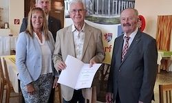 Drei Personen stehen vor einem Banner und halten ein Stück Papier in der Hand. Ein Mann in der Mitte hält ein Papier mit einem Design. Die anderen lächeln und schauen in die Kamera. Dahinter steht ein Tisch mit Stühlen.