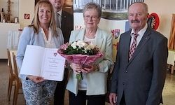 Drei Personen stehen in einer Innenumgebung; eine Frau hält ein Zertifikat und einen Blumenstrauß, eine andere Frau hält einen Blumenstrauß und ein Mann steht neben ihnen in einem Anzug.